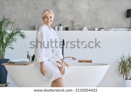 Wideshot of happy smiling beautiful middle aged woman in bathrobe at luxury spa hotel sitting on bath tube. Advertising of bodycare spa procedures antiage recreation skin care salon concept. Royalty-Free Stock Photo #2135054433