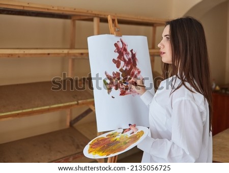 A young brunette artist in a white shirt paints on canvas in her studio. Next to her is an easel, paintings and various artistic techniques.