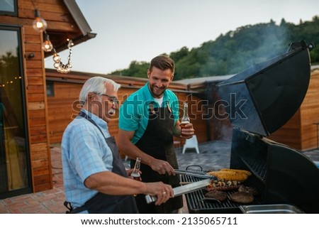 Pensioner and his son wearing aprons, holding bottles of beer and roasting meat on barbecue in the backyard Royalty-Free Stock Photo #2135065711