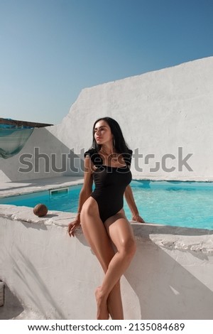 vacation mood. brunette woman in black glamour swimsuit sits on the swimming pool near blue water with coconut and smiling cute near white wall background. greek villa. lifestyle concept, free space