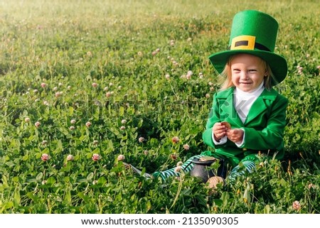 St. Patrick's Day. A girl in a green suit is sitting in a large clearing of clover, holding a pot with gold coins in her hands. copyspase. Happy joyful toddler baby