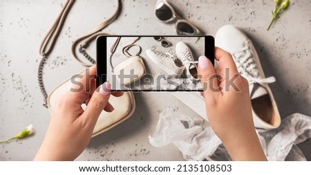Woman taking photo of white leather sneakers shoes, handbag and sunglasses with smartphone. Blogger, influencer or stylist capturing spring fashion accessories for social media. Grey background.