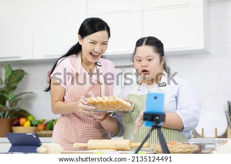 mother and down syndrome teenage girl or her daughter showing bread on basket and live streaming online via smartphone on tripod in a kitchen