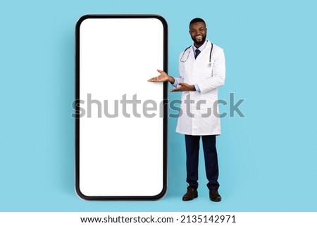 Online Offer. Black Smiling Doctor In White Robe Pointing At Big Blank Smartphone With Empty Screen For Mockup, African American Therapist Showing Copy Space For Mobile App Design Or Advertisement Royalty-Free Stock Photo #2135142971
