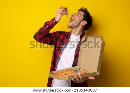 Excited Young Guy Enjoying Pizza Holding And Biting Tasty Slice Posing With Open Mouth Holding Carton Box On Yellow Orange Studio. Junk Food Lover Eating Italian Pizza. Unhealthy Nutrition Cheat Meal Royalty-Free Stock Photo #2135143007