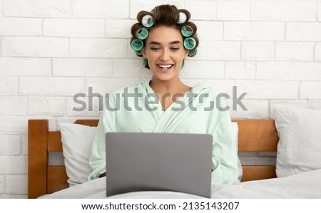 Happy Glamorous Lady Using Laptop Computer Working Online Browsing Internet Sitting Wearing Dressing Gown Curling Hair Sitting In Bed In Modern Bedroom At Home. Distance Job And Freelance Career Royalty-Free Stock Photo #2135143207