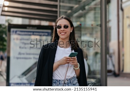 Outdoor portrait of pretty stylish girl in dark jacket in black glasses is scrolling smartphone and smiling while walking in the city in sunny warm day