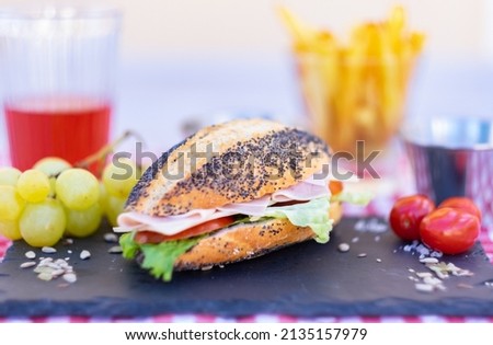 Poppy seed sandwich with vegetables and cooked ham on a black stone cutting board. Light and healthy snack, French fries and a glass of wine in the background.