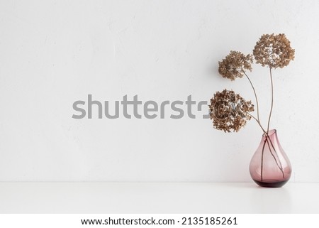White desk with pink glass vase with a decorative dried branches, flower against white wall.	
