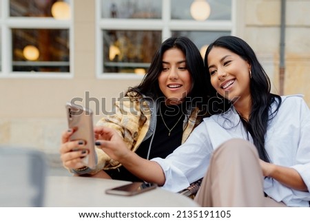 two hispanic sisters taking a picture with their smart phone in the street. Young latin girls using a mobile phone and having fun happily.