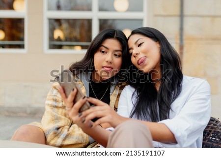 Two Latin women friends take pictures of themselves sticking out their tongues and gesturing with their smartphone. Latin sisters having good times together in the street.