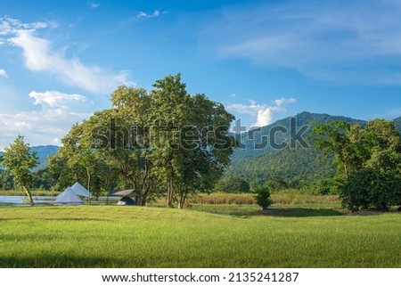 Traveling and camping concept.  Camping tent in forest near lake with grassland and blue sky at the morning. natural landscape in Kanchanaburi province, Thailand at summer time so warm and relax. Royalty-Free Stock Photo #2135241287