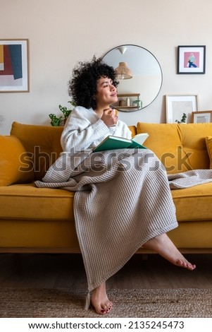 Pensive happy African american woman reading a book at home sitting on sofa relaxing drinking coffee. Vertical image. Royalty-Free Stock Photo #2135245473