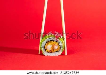 Single sushi roll with shrimp, avocado and flying fish caviar on red background with wooden sticks. Creative image on color backdrop with long shadow. Royalty-Free Stock Photo #2135267789