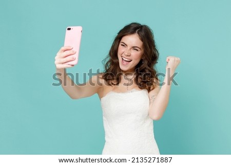 Happy bride young woman 20s in white wedding dress doing selfie shot on mobile phone doing winner gesture isolated on blue turquoise background studio portrait. Ceremony celebration party concept