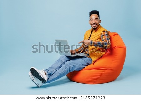 Full size body length young black man 20s wears yellow waistcoat shirt sit in bag chair hold using work pointing on laptop pc computer isolated on plain pastel light blue background studio portrait