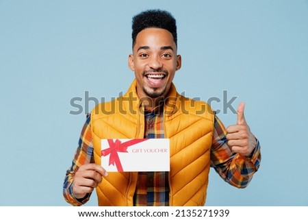 Excited young black man 20s wears yellow waistcoat shirt hold gift certificate coupon voucher card for store show thumb up like gesture isolated on plain pastel light blue background studio portrait