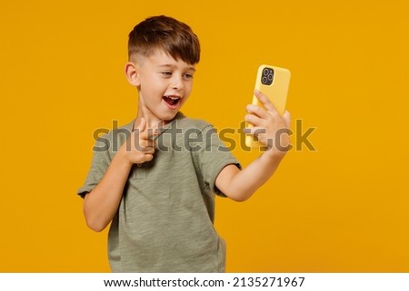 Little small smiling boy 6-7 years old in green t-shirt doing selfie shot on mobile cell phone point finger on camera isolated on plain yellow background. Mother's Day love family lifestyle concept