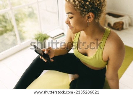 Close-up picture of delighted curly blonde short hair girl with perfect dark skin sitting in white room in fitness clothes wearing piercing typing on her phone with screen space for your advertisement
