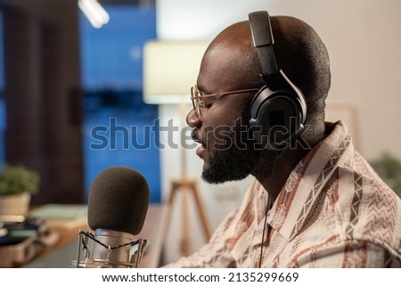 Side view of young black man in casual shirt, headphones and eyeglasses singing and recording his songs in studio or at home Royalty-Free Stock Photo #2135299669