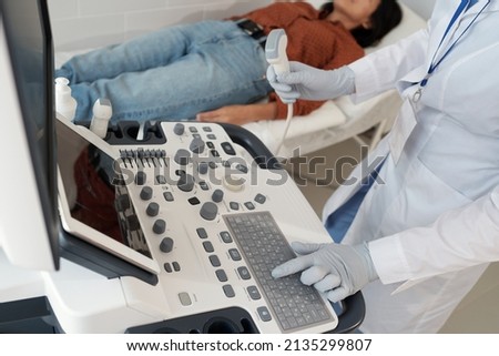 Gloved hand of female clinician pressing button on keypad of ultrasound machine while going to examine young patient on couch Royalty-Free Stock Photo #2135299807