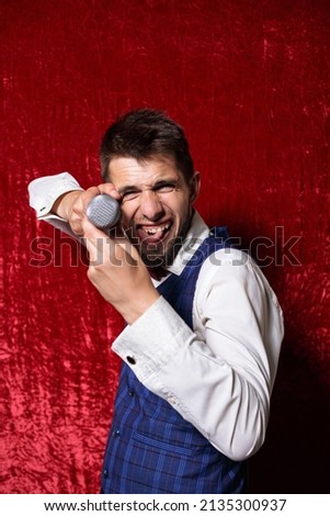 Excited showman with mic and in suit making funny face and performing while looking at camera on red background 