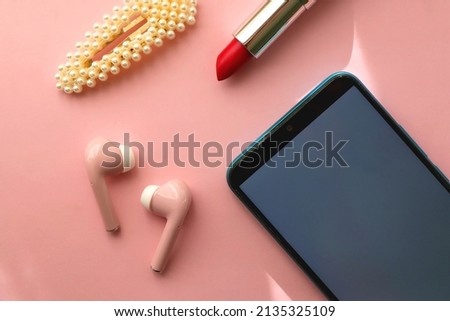 Mobile phone, pink wireless earphones, red lipstick and pearl hair clip on pastel pink background. Flat lay.