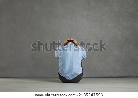 Back view studio portrait of man sitting on floor facing grey stone wall, holding head, trying to hide true emotions, feeling desperate, distressed, frustrated, hopeless, unable to cope with problem Royalty-Free Stock Photo #2135347553