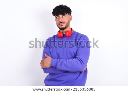 Pleased young arab man with curly hair wearing purple sweatshirt over white background keeps hands crossed over chest looks happily aside