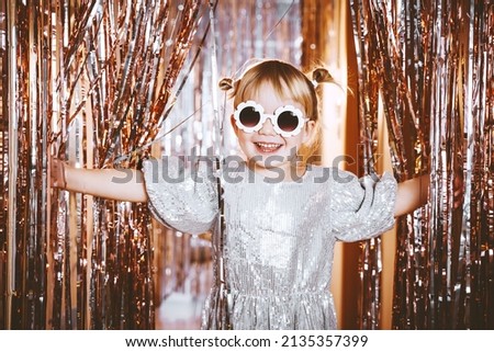 Happy little stylish girl in shiny dress having fun. Festive background with foil curtain decorations for kids birthday or fancy dress party, disco music or New Year. Celebration and Holiday concept. Royalty-Free Stock Photo #2135357399