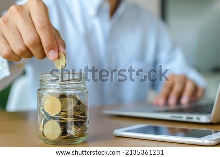 Young businessman wearing blue shirt holding coins putting in glass on desk with copy space. Concept saving money for education and retirement.