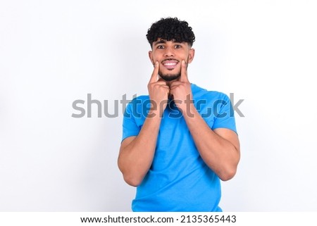 Happy young arab man with curly hair wearing blue t-shirt over white background with toothy smile, keeps index fingers near mouth, fingers pointing and forcing cheerful smile