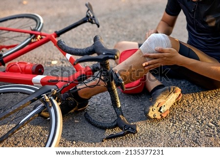 Accident man in sports concept. biker fall from bike and wrapping leg in bandage. cropped injured young male bycyclist in black sportswear has an accident on bike, red bike on road next to him Royalty-Free Stock Photo #2135377231