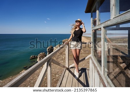 young woman traveler in a straw hat and dark glasses stands on the balcony of a wooden house against the backdrop of a beautiful seascape. her long hair flutters in the wind