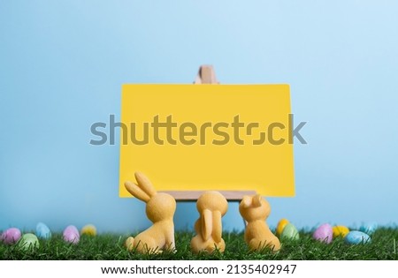 Three easter bunnies sitting on the grass rear view among the painted eggs and look at blank canvas on an easel. Easter advertising banner