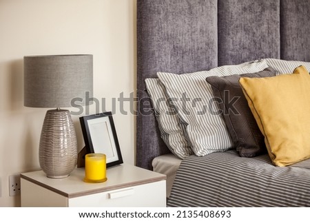 Modern bedroom bed detail with fabric headboard, pillows, cushions and sidetable drawer with lamp, candle and picture frame