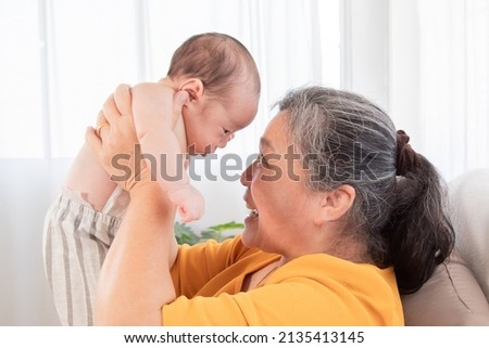 Side view grandmother and newborn baby play together at home, Grandma holding adorable infant in air and looking with love and tender. Spend time together. Senior women making funny faces to toddler.