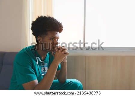 Shot of a young doctor looking distressed. Tired exhausted male african scrub nurse wears green uniform gloves sits on hospital. Depressed sad black ethic doctor feels burnout stress Royalty-Free Stock Photo #2135438973