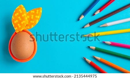 Egg with rabbit ears on a blue background with colorful wooden crayons. Copy space. Flat lay. Easter concept.