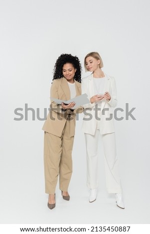 Positive african american businesswoman holding paper folder near colleague with smartphone on grey background