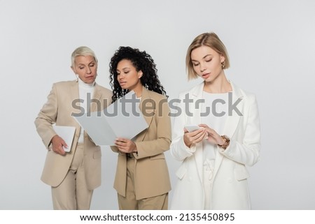 Blonde businesswoman using smartphone near interracial colleagues with paper folder isolated on grey