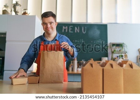 Cheerful young Down Syndrome waiter working in take away restaurant, social inclusion concept. Royalty-Free Stock Photo #2135454107