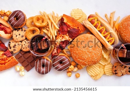 Junk food table scene scattered over a white marble background. Collection of take out and fast foods. Pizza, hamburgers, french fries, chips, hot dogs, sweets. Top view. Royalty-Free Stock Photo #2135456615