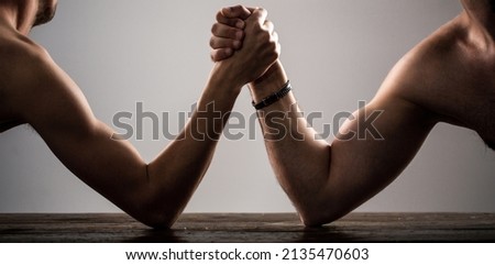 Heavily muscled bearded man arm wrestling a puny weak man. Arms wrestling thin hand, big strong arm in studio. Two man's hands clasped arm wrestling, strong and weak, unequal match. Arm wrestling.