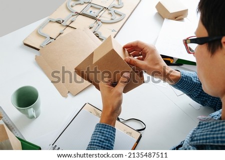 Young box designer looking at the folding of the product. Checking alignment. Sitting behind the desk, wearing glasses.