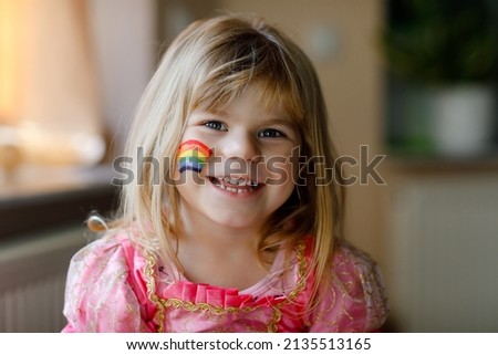 Portrait of cute little toddler girl with painted rainbow with colorful colors on face. Happy child with sign for diversity and peace in the world.