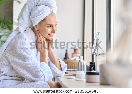 Attractive mid age older adult 50 years old blonde woman wears bathrobe and towel in bathroom touching face, looking at mirror doing daily beauty routine. Skin care treatment concept. Royalty-Free Stock Photo #2135514545