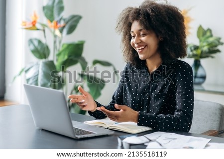 Successful positive young adult woman african woman freelancer, manager, CEO, sitting in office at laptop, talking on video call with client or employees, discussing business strategy, gesturing,smile Royalty-Free Stock Photo #2135518119