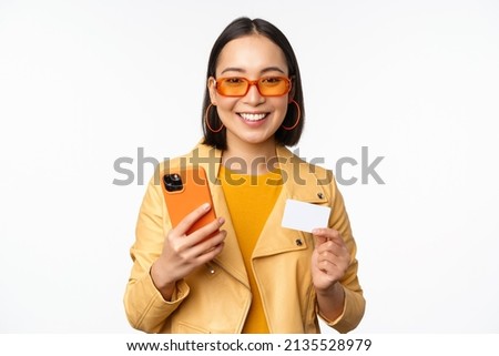 Online shopping. Stylish asian female model in sunglasses, holding credit card and mobile phone, smiling happy, standing over white background