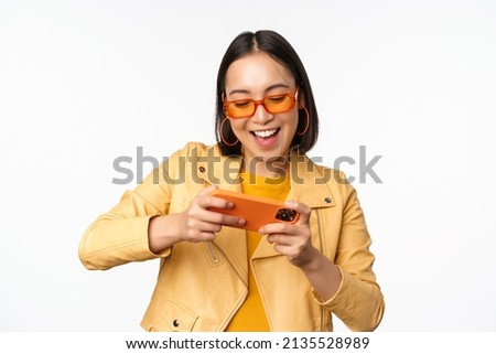 Stylish korean girl in sunglasses, playing mobile video game, laughing and smiling while using smartphone, standing over white background